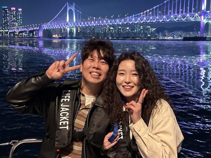 Two guests smiling on the yacht with the lit-up Gwangan Bridge behind